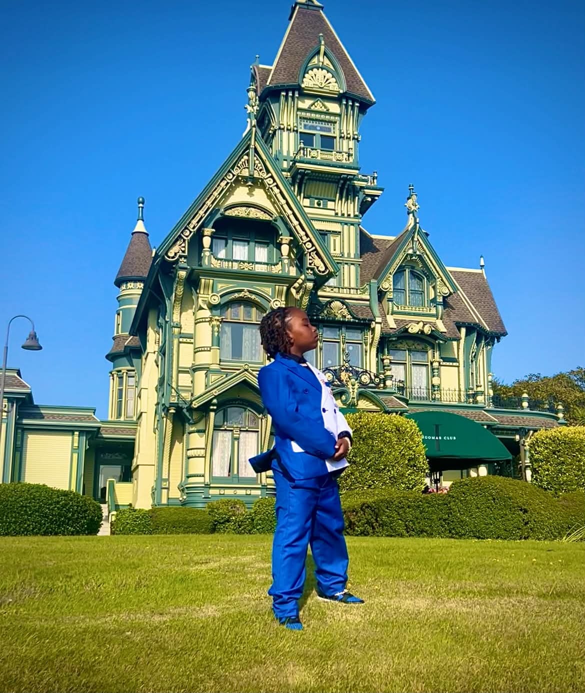 Child in a blue suit standing before an ornate Victorian home